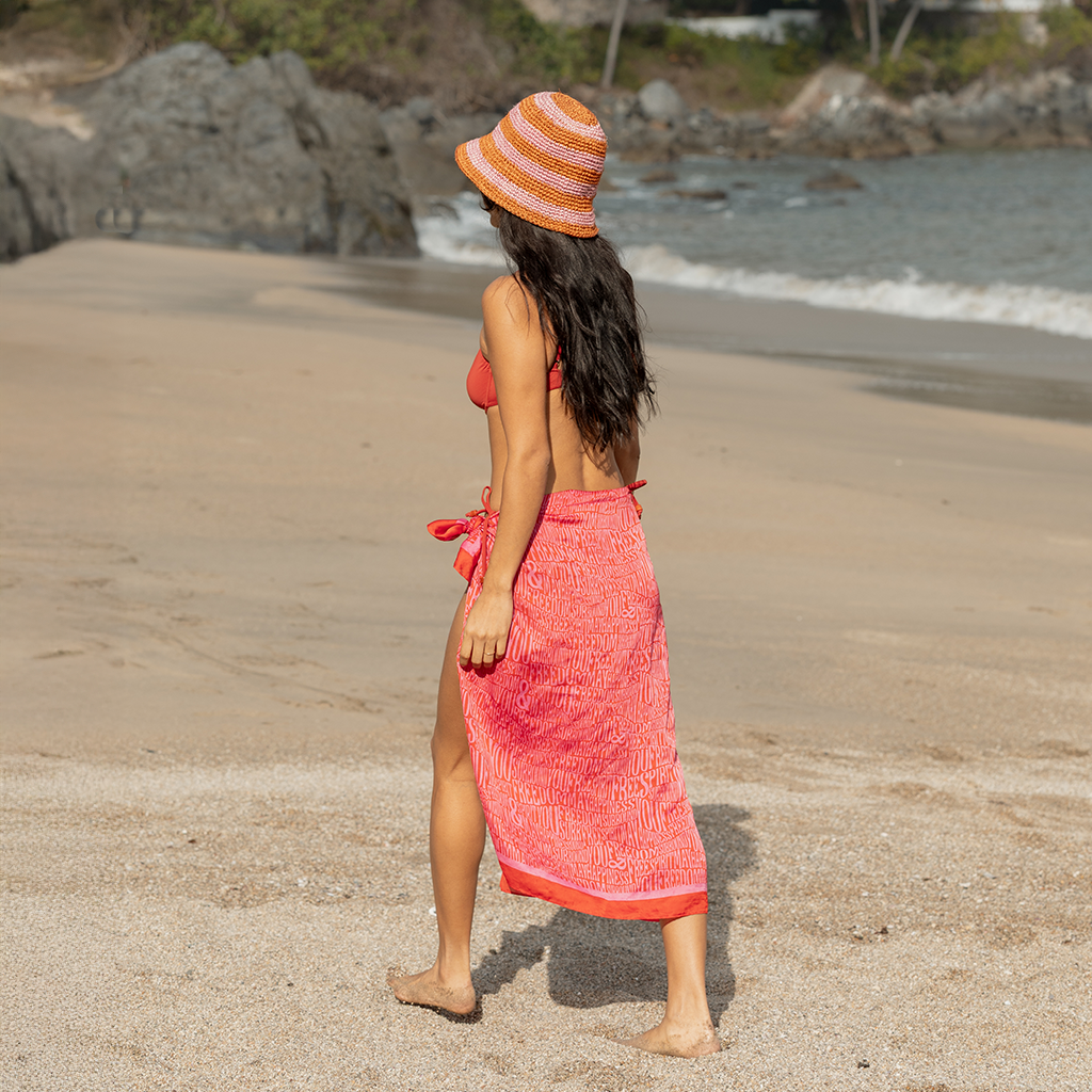 Woman in a red bikini and pink sarong walking on a beach wearing a striped hat