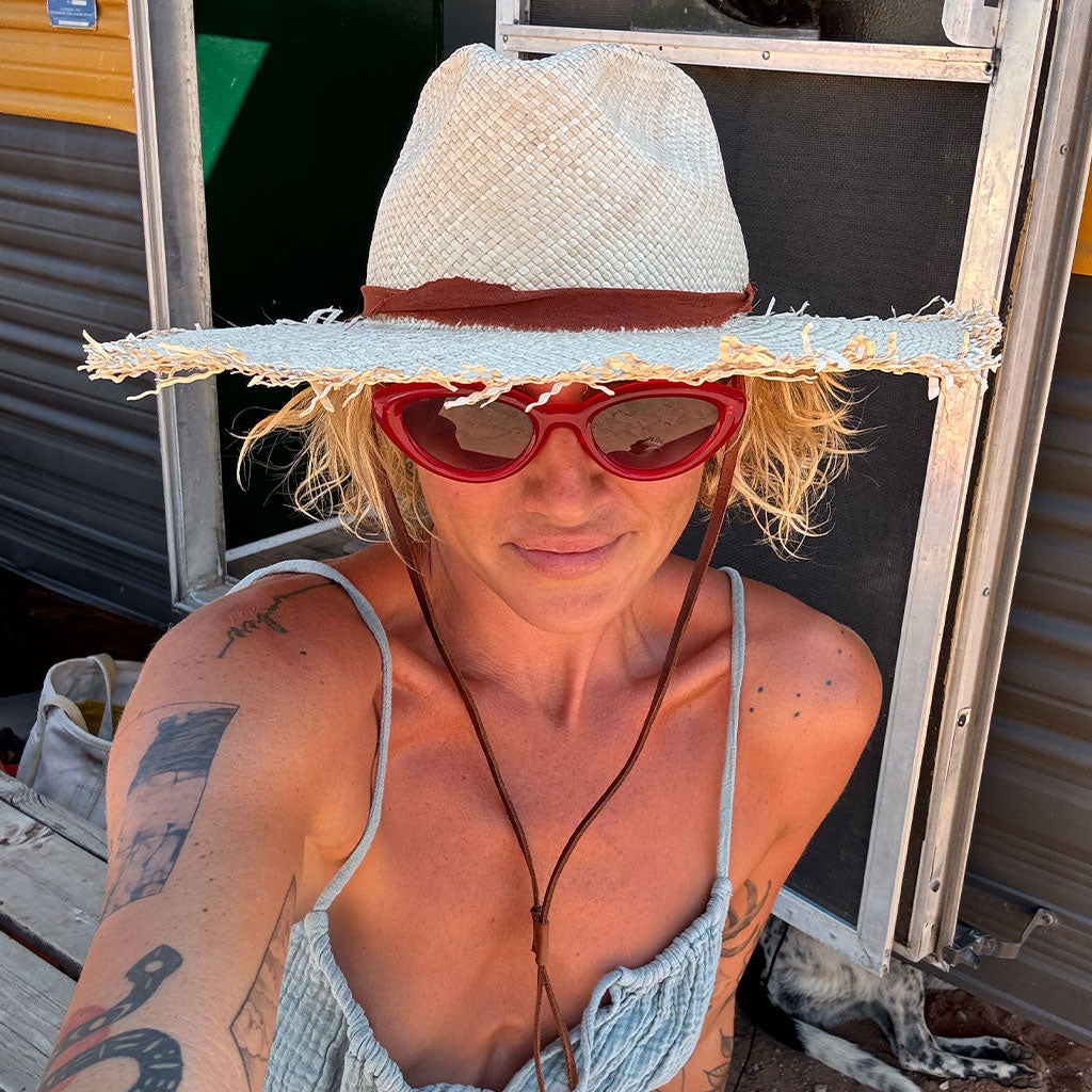 Person wearing a straw hat and red sunglasses with tattoos visible on arms, standing in front of a metal structure.